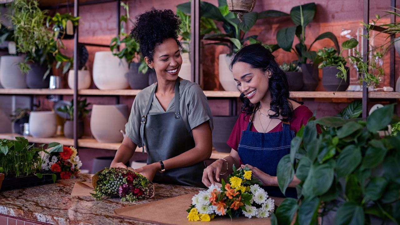 Duas floristas jovens felizes e sorridentes trabalhando juntas em uma floricultura. Florista conversando e fazendo um lindo buquê de flores junto com sua colega negra. Lindas floristas arrumando o buquê.